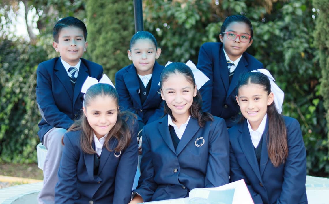Seis niños en uniforme escolar azul oscuro posando al aire libre con hojas verdes de fondo.