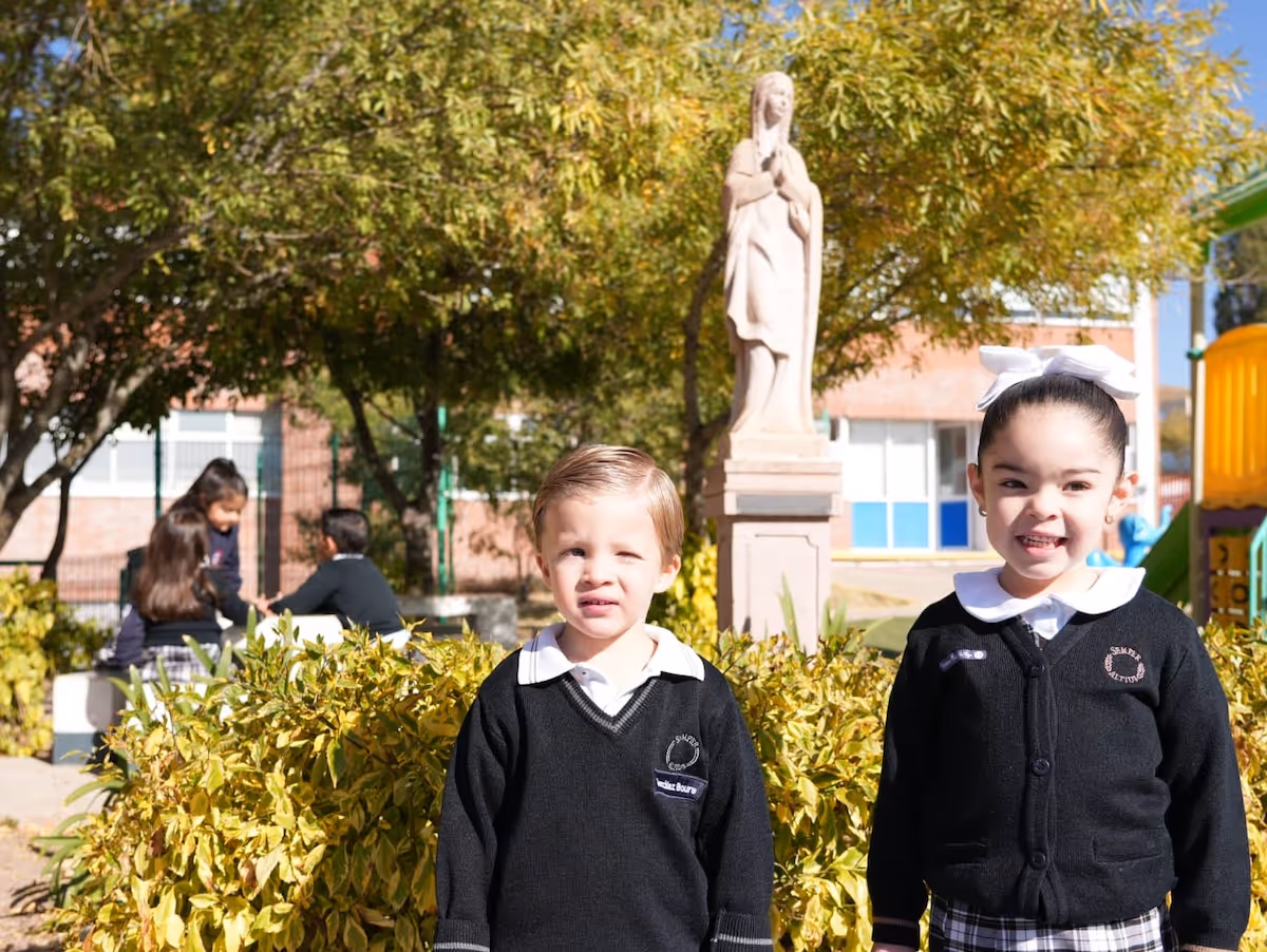 Dos niños con uniforme escolar negro posan al aire libre frente a un seto y una estatua en un día soleado.