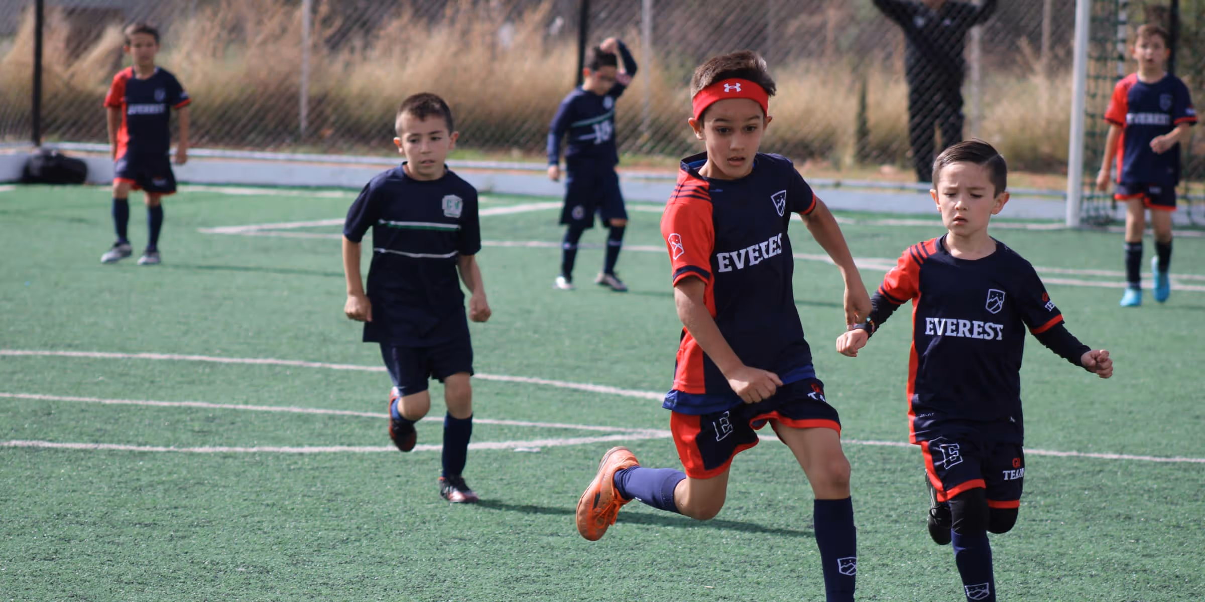 Niños jugando fútbol en un campo con uniforme azul y rojo, dos niños adelante corriendo juntos.